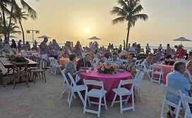 People seated at tables and standing on the beach at sunset